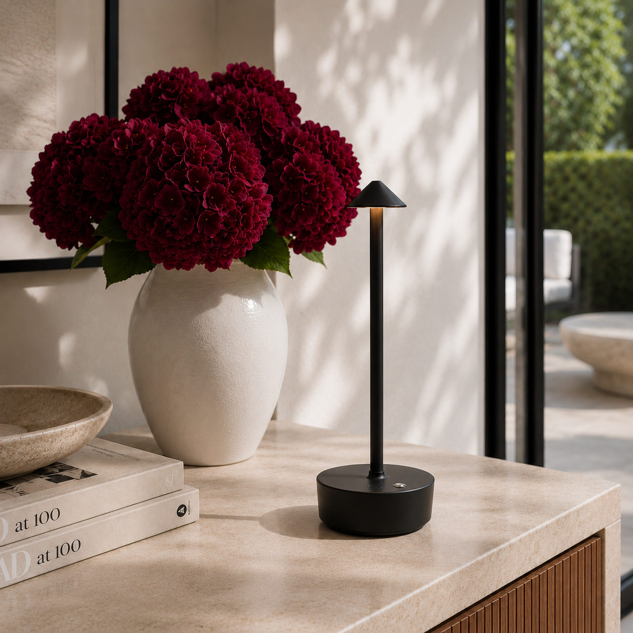 Black halo light with with burgundy flowers and books on a kitchen counter.  The lamp is cordless, portable, rechargeable and weatherproof.  Available in black, gold and brown.