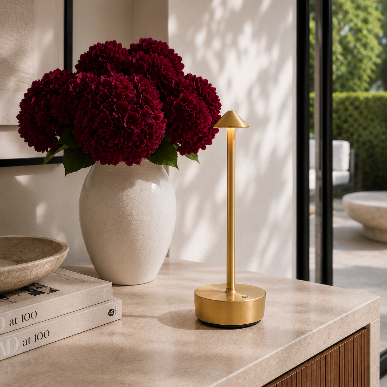 Gold halo light with with burgundy flowers and books on a kitchen counter.  The lamp is cordless, portable, rechargeable and weatherproof.  Available in black, gold and brown.