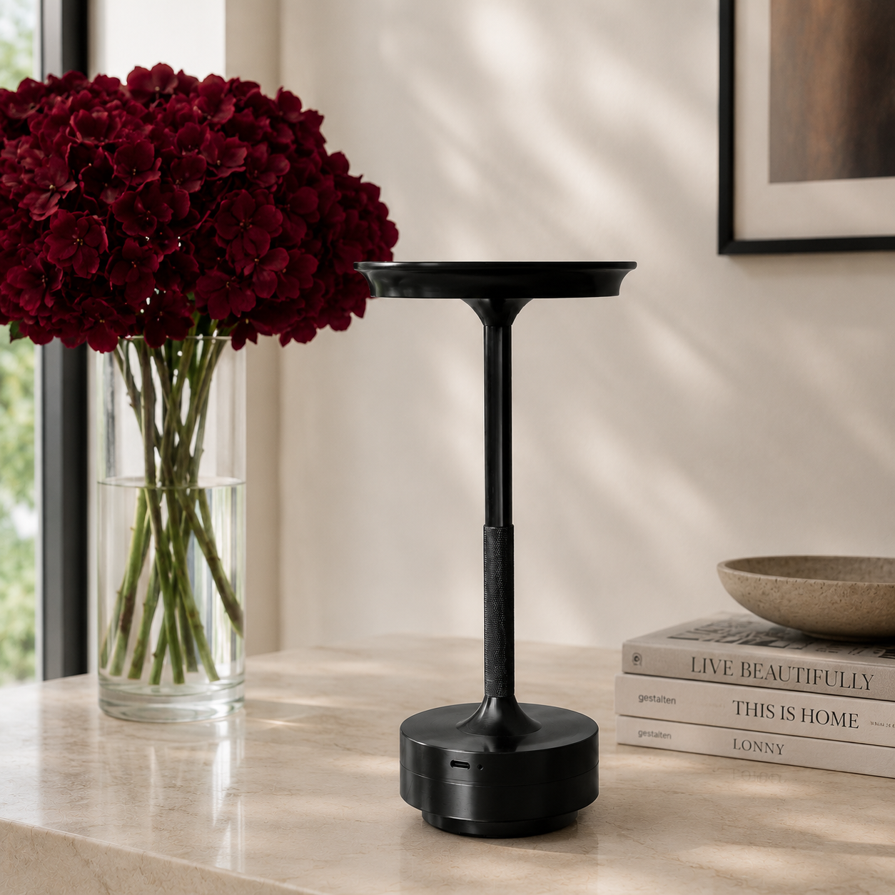 A black cordless table lamp with a circular base and wide top, styled on a light marble tabletop next to a vase of deep red flowers and a stack of books. It is portable, rechargeable via USB-C and is weatherproof. 