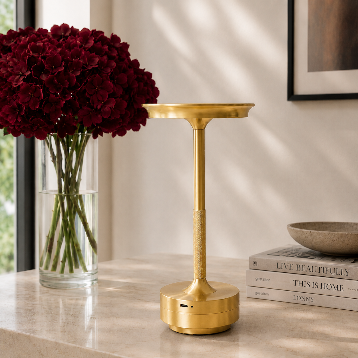 A gold 
cordless table lamp with a circular base and wide top, styled on a light marble tabletop next to a vase of deep red flowers and a stack of books. It is portable, rechargeable via USB-C and is weatherproof. 