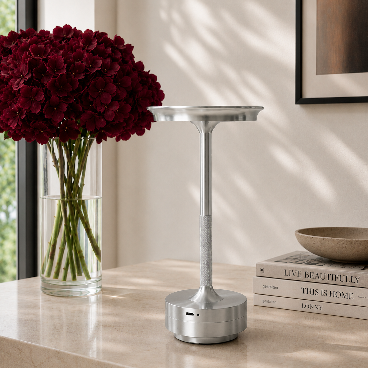A silver cordless table lamp with a circular base and wide top, styled on a light marble tabletop next to a vase of deep red flowers and a stack of books. It is portable, rechargeable via USB-C and is weatherproof. 