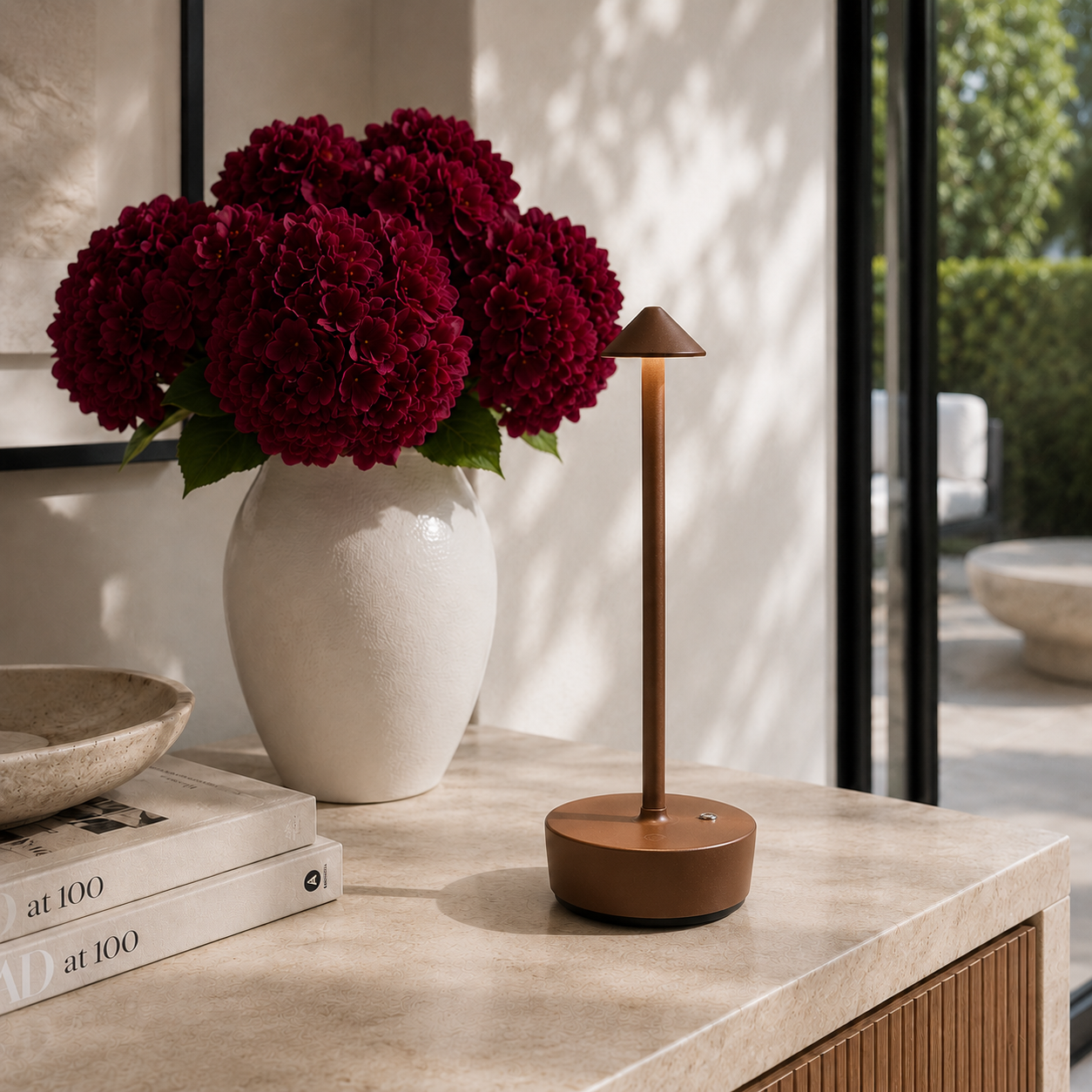 Brown halo light with with burgundy flowers and books on a kitchen counter.  The lamp is cordless, portable, rechargeable and weatherproof.  Available in black, gold and brown.