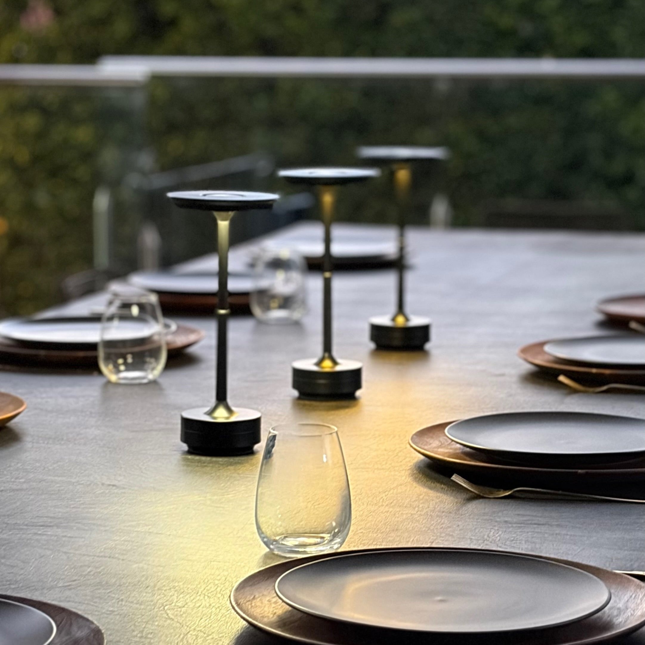 Outdoor dining table set with plates, glasses, and 3 lamps on a reflective surface. The lights are cordless, portable, rechargeable and weatherproof.