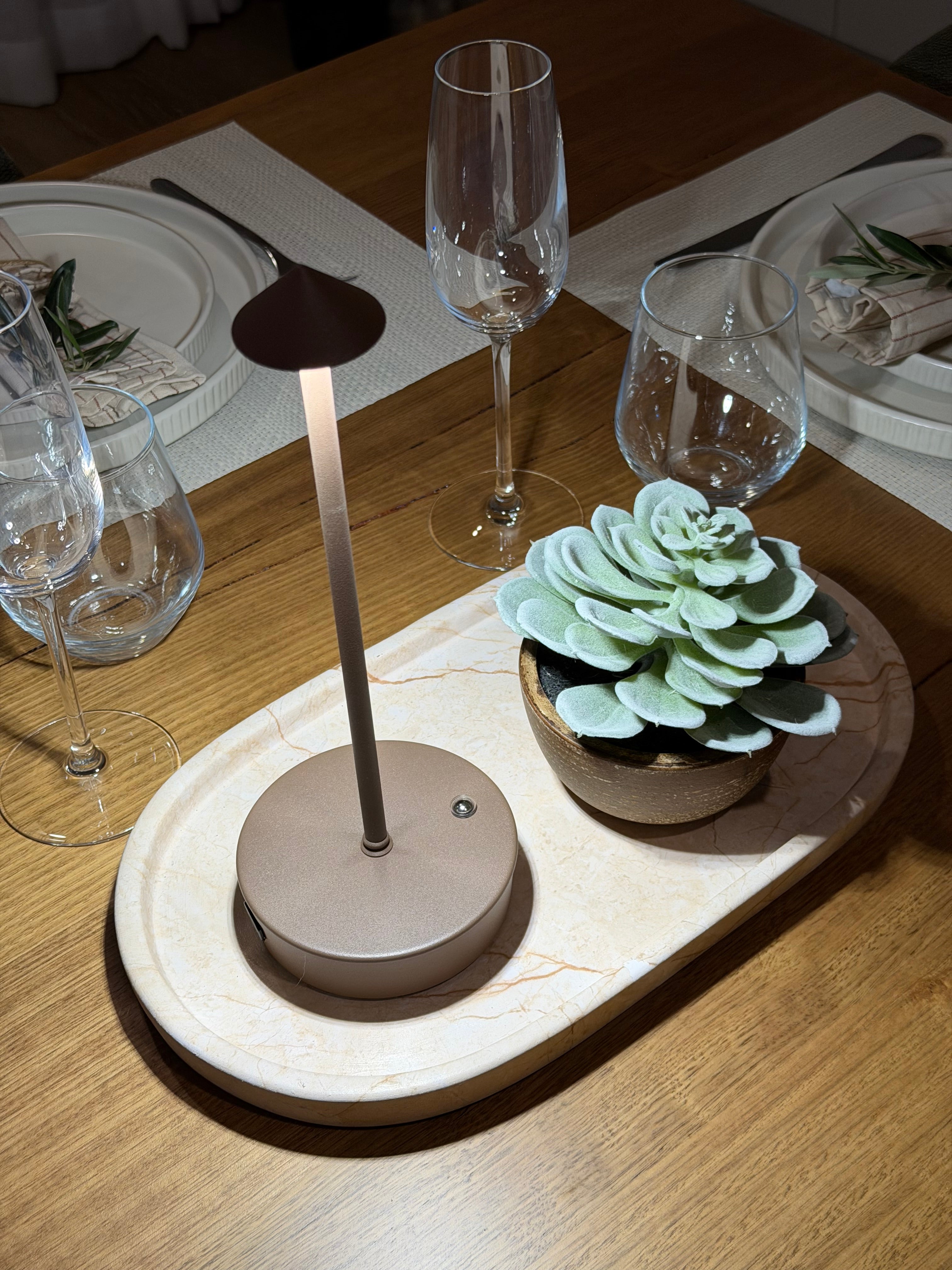 Brown halo light with decorative table setting with wine glasses, a plant, and a lamp on a wooden surface.